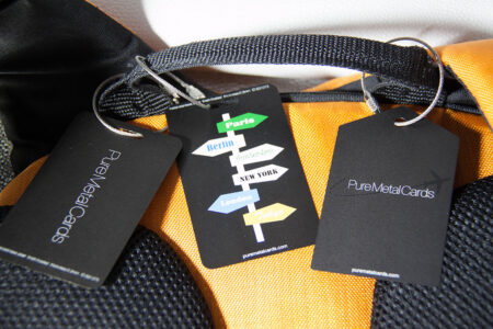 Orange luggage with black metal luggage tags attached. Tags read "PureMetalCards.