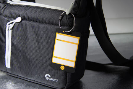 Black camera bag with attached metal luggage tag. The tag has space to fill in name, address, and phone number.