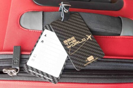 Sleek carbon fiber luggage tag attached to a red suitcase. "See the World" is visible on the tag.