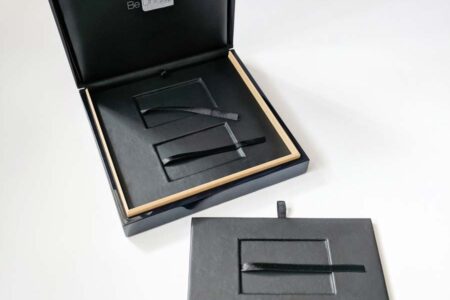 Open black luxury wood presentation case with compartments and black ribbons. "Be Unique" logo visible.