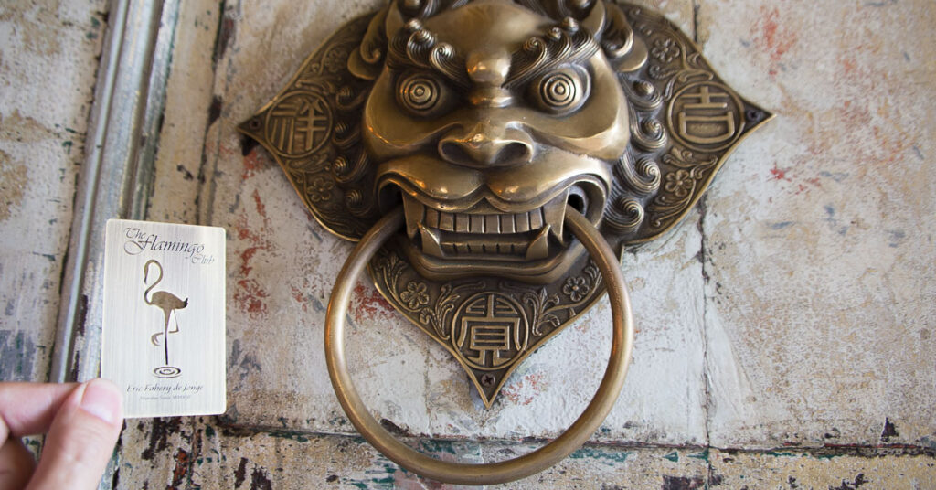 Metal business card of The Flamingo Club held against ornate brass door knocker. Prestige.