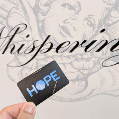Hand holding a metal card with "HOPE IS..." against a background with the word "Whispering". Reflecting brand identity.
