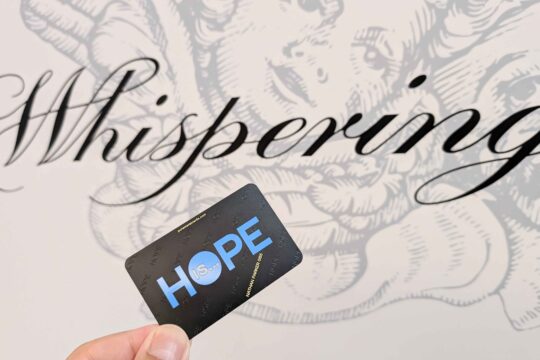 Hand holding a metal card with "HOPE IS..." against a background with the word "Whispering". Reflecting brand identity.