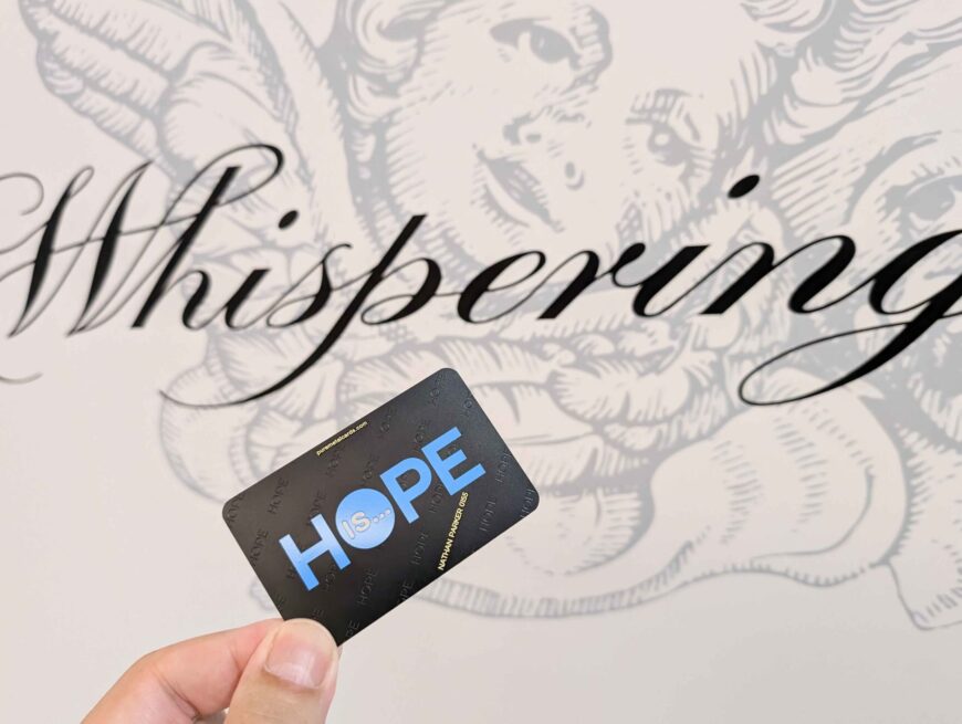Hand holding a metal card with "HOPE IS..." against a background with the word "Whispering". Reflecting brand identity.