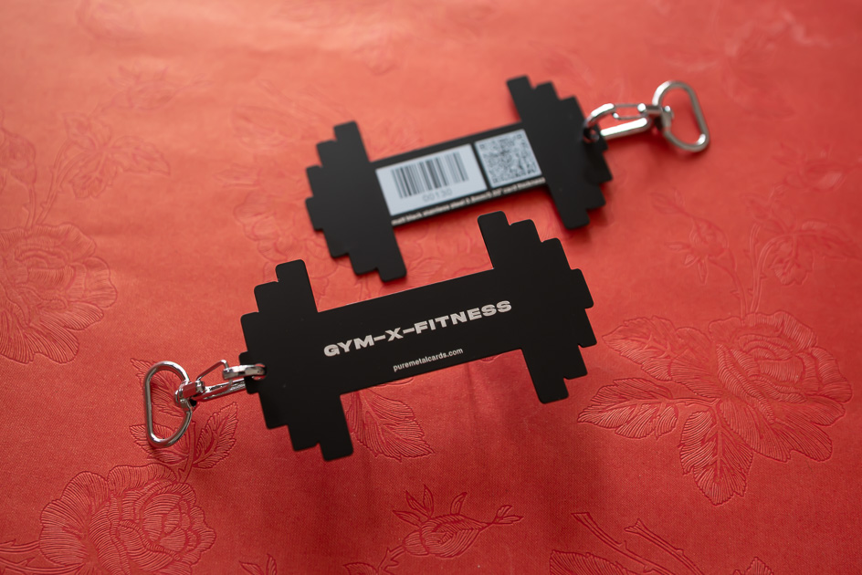 Two black dumbbell-shaped metal membership cards with keyrings. GYM-X-FITNESS is printed on one.