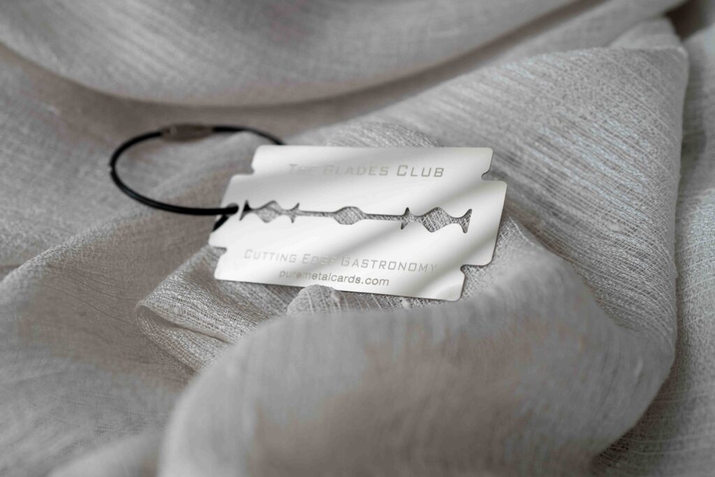 Metal member card shaped like a razor blade on textured fabric. Text: "The Blades Club. Pure Metal Cards
