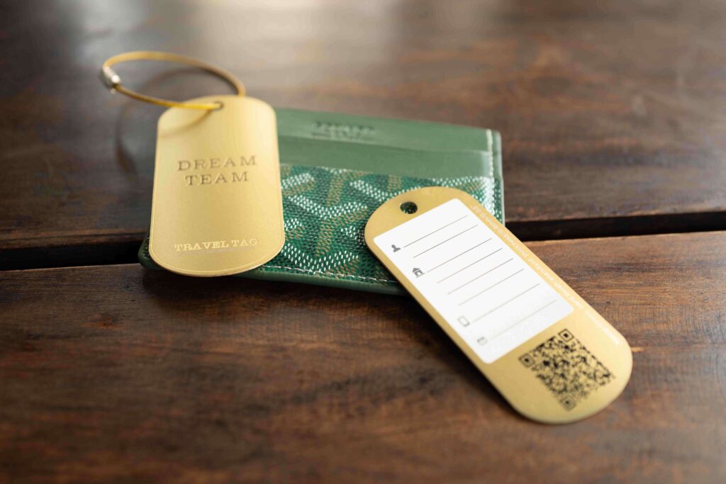 Metal luggage tags with "Dream Team Travel Tag" text next to a green wallet on a wooden surface. Pure Metal Cards